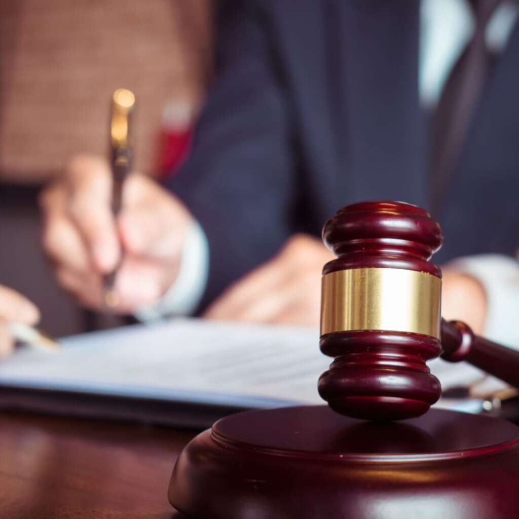Gavel in view on a desk with a person in a courtroom writing on a. document in the background about trade secrets.